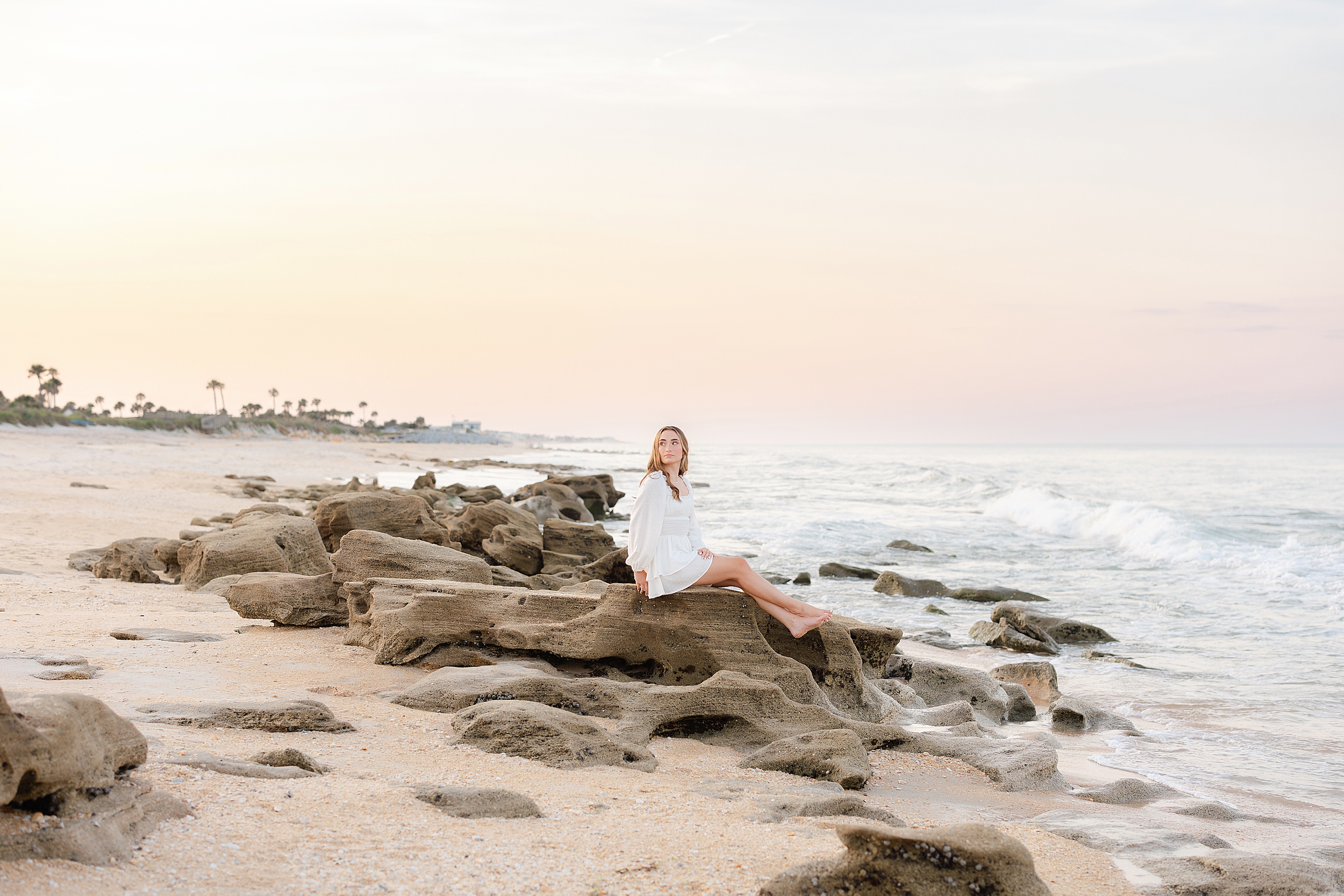 Grad portrait of young woman in white dress sitting on the rocks in St. Augustine Beach at sunset.