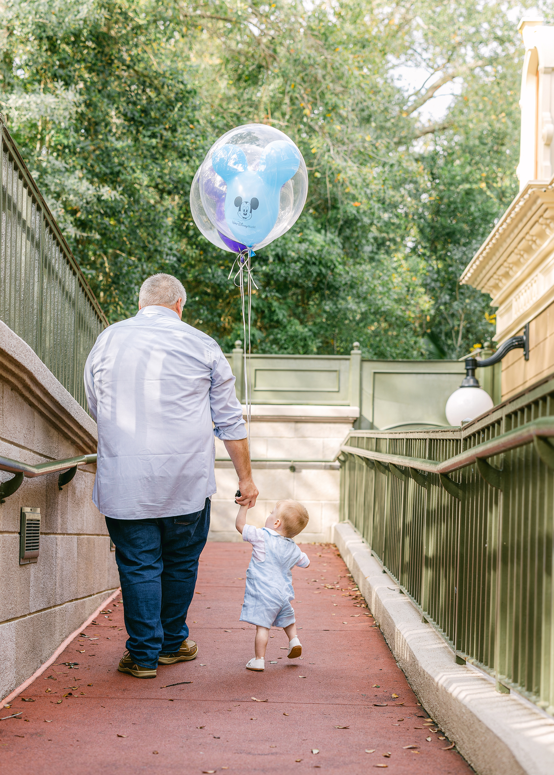 A baby boy dressed in blue and white holds a Mickey Balloon as he looks up at his father.