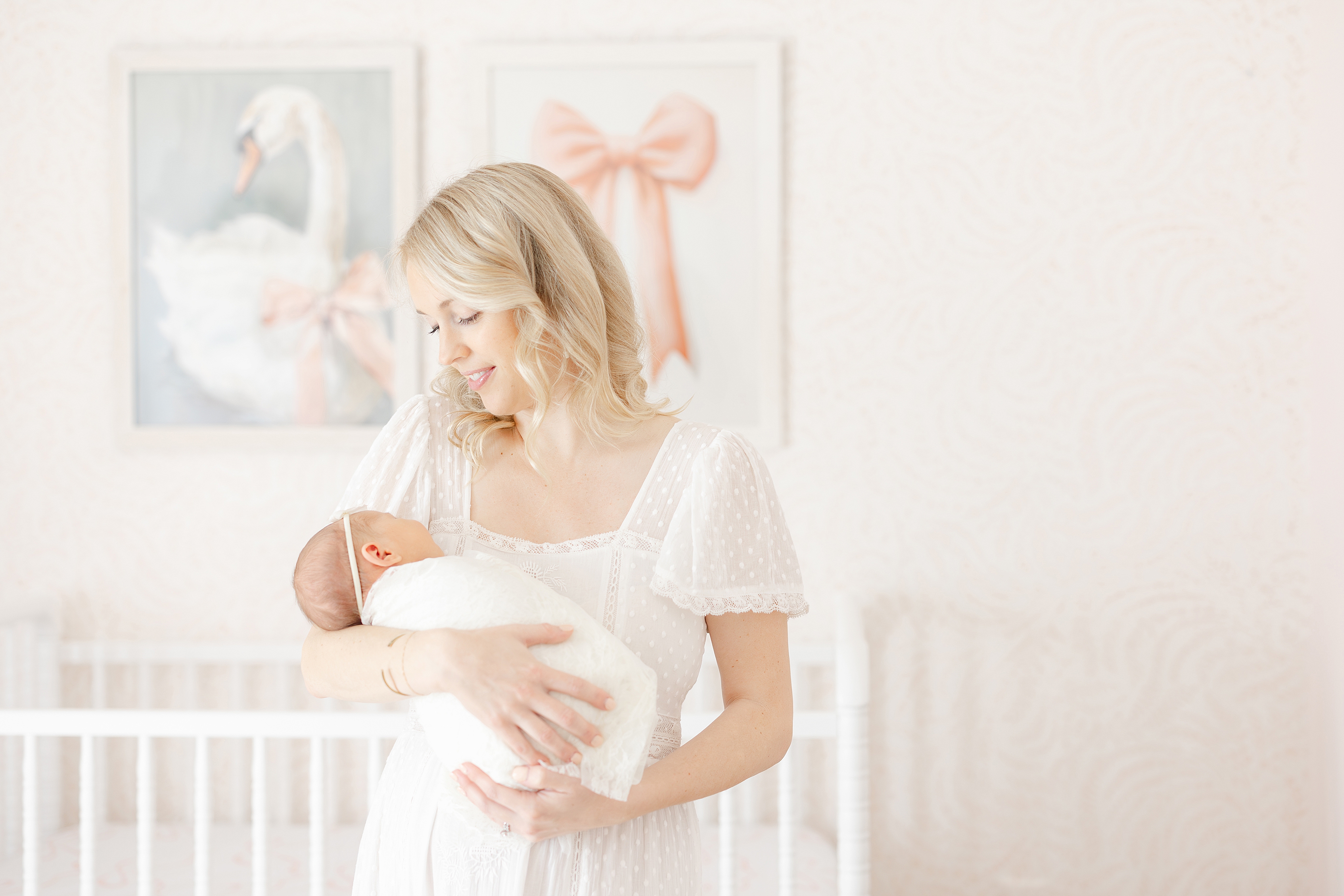 Woman in white lace dress holding a newborn baby gilr in a pink and white nursery.