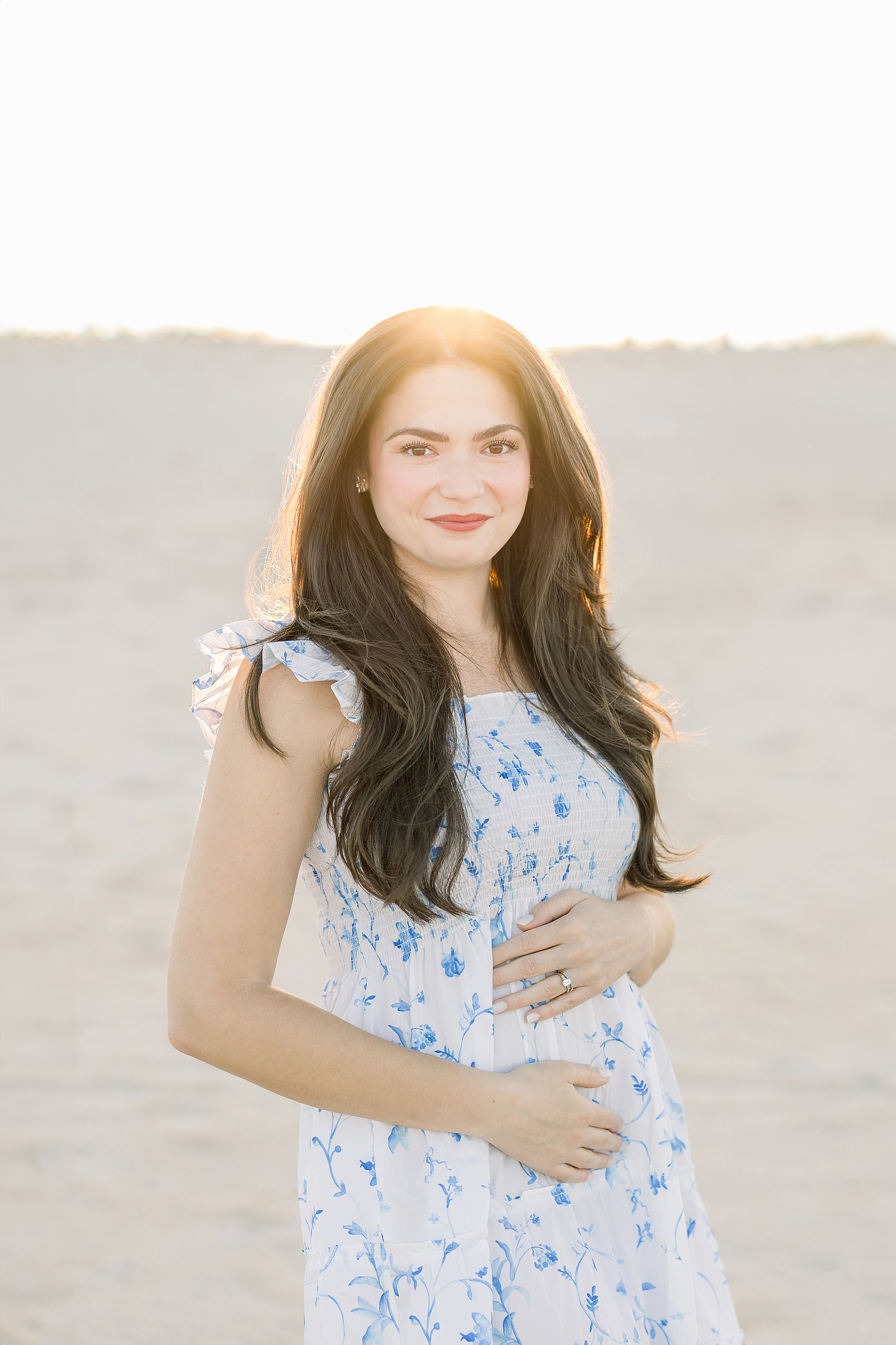 A pregnancy announcement portrait of a woman in a blue and white dress on the beach at sunset.