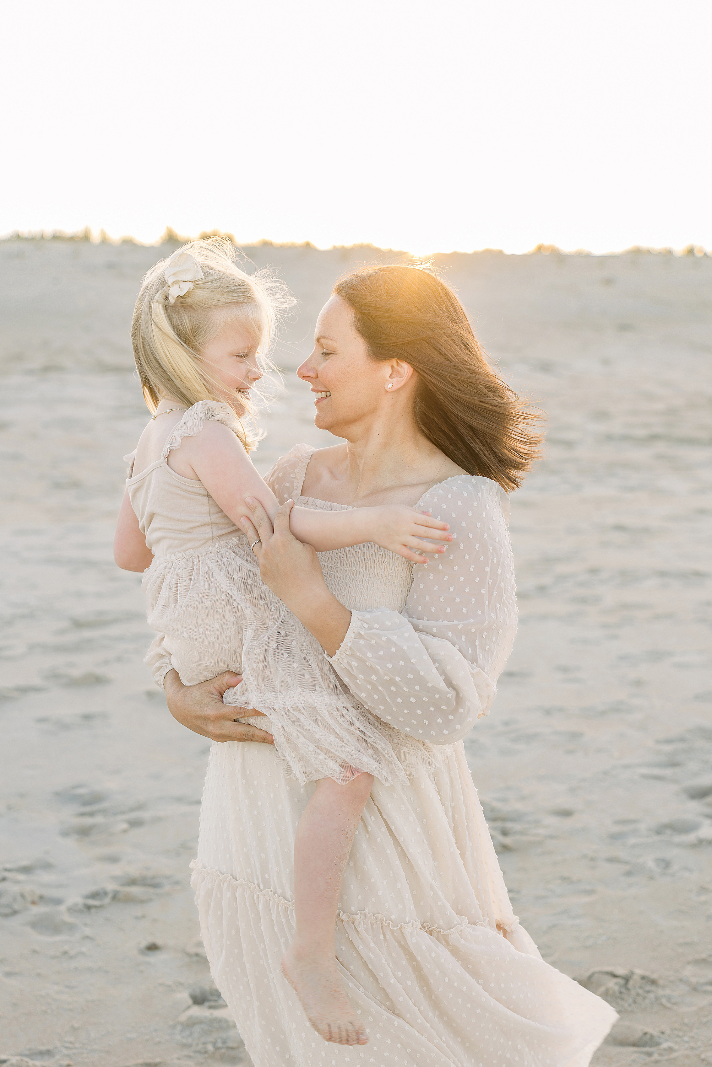 A mother and her little girl dressed in cream tulle dresses hold each other on the beach at sunset.