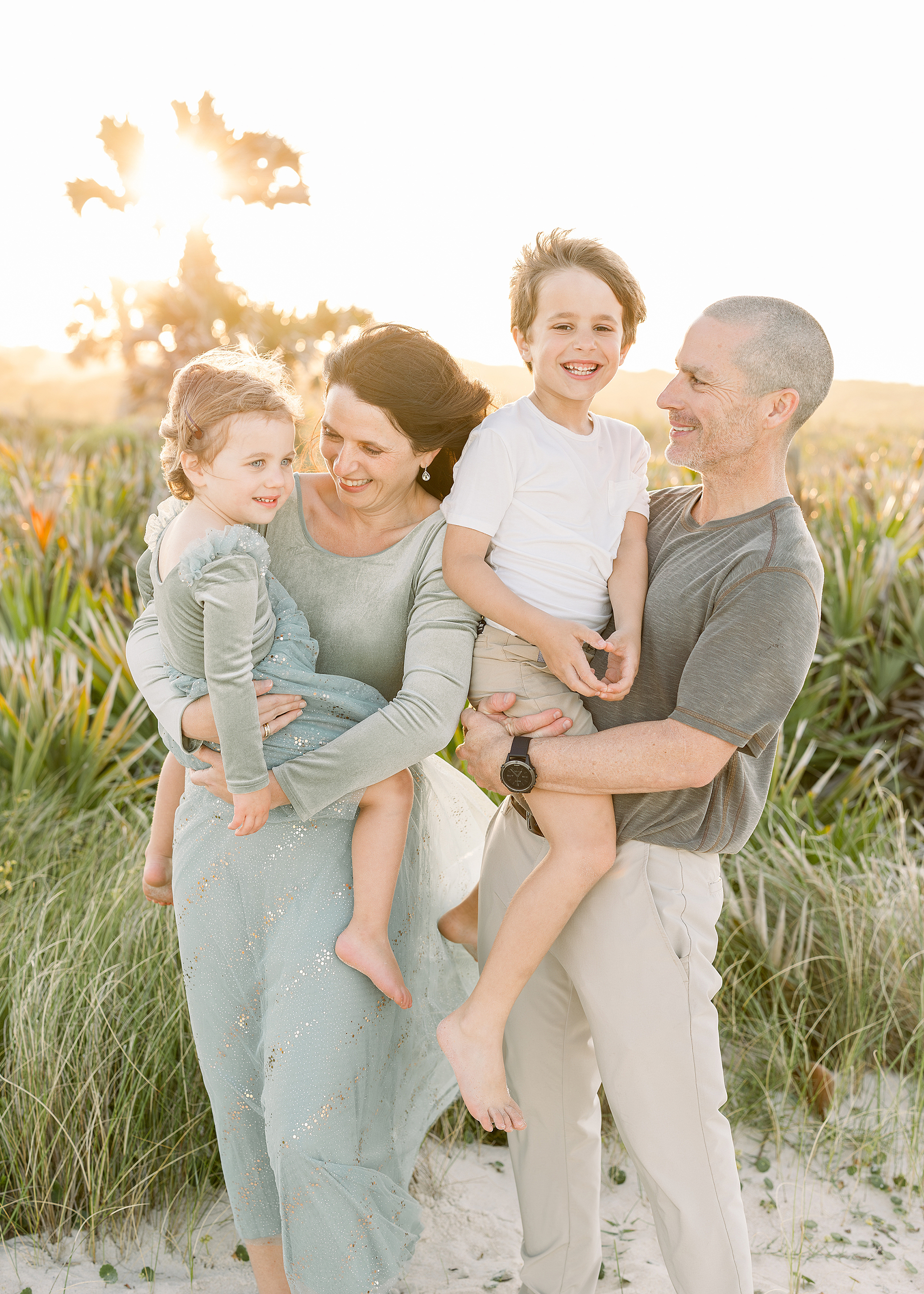 A family of four wearing sage green and neutral colors laugh together with each other on the beach at sunset.