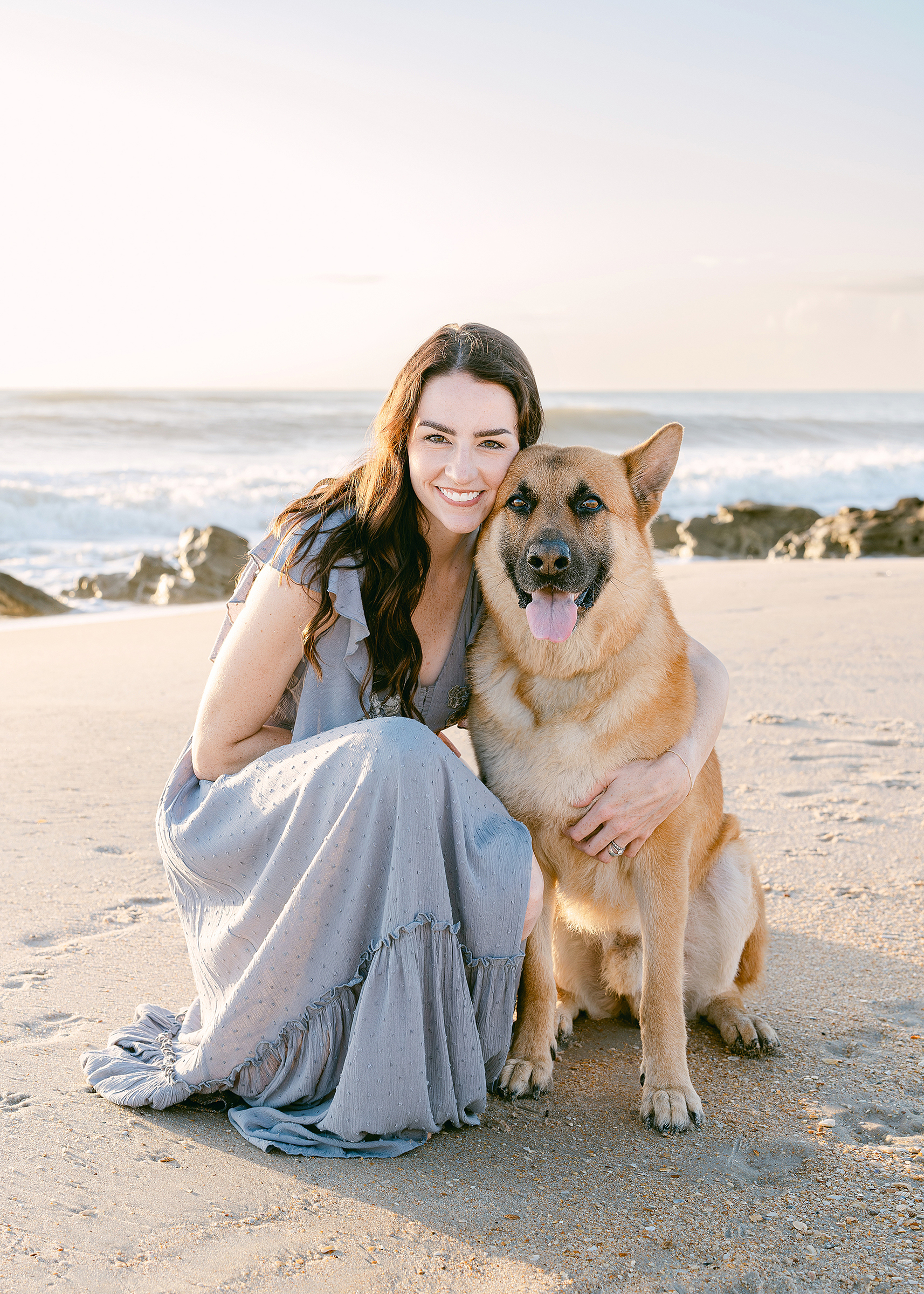 A woman in a lavender dress kneels down on the sandy beach with her dog at sunrise.