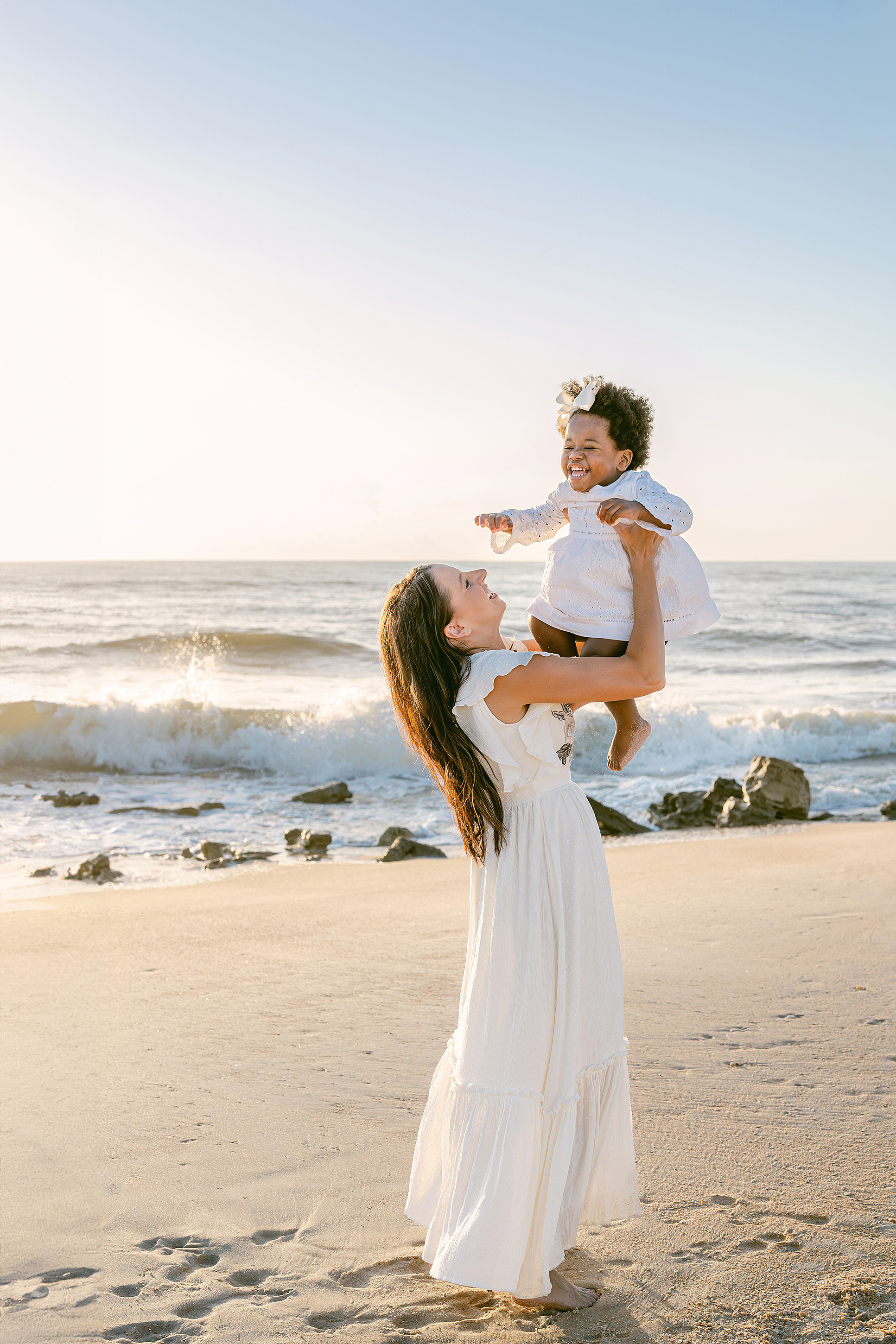 A light and airy sunrise motherhood portrait of a woman throwing a little black girl in a white dress at the beach.