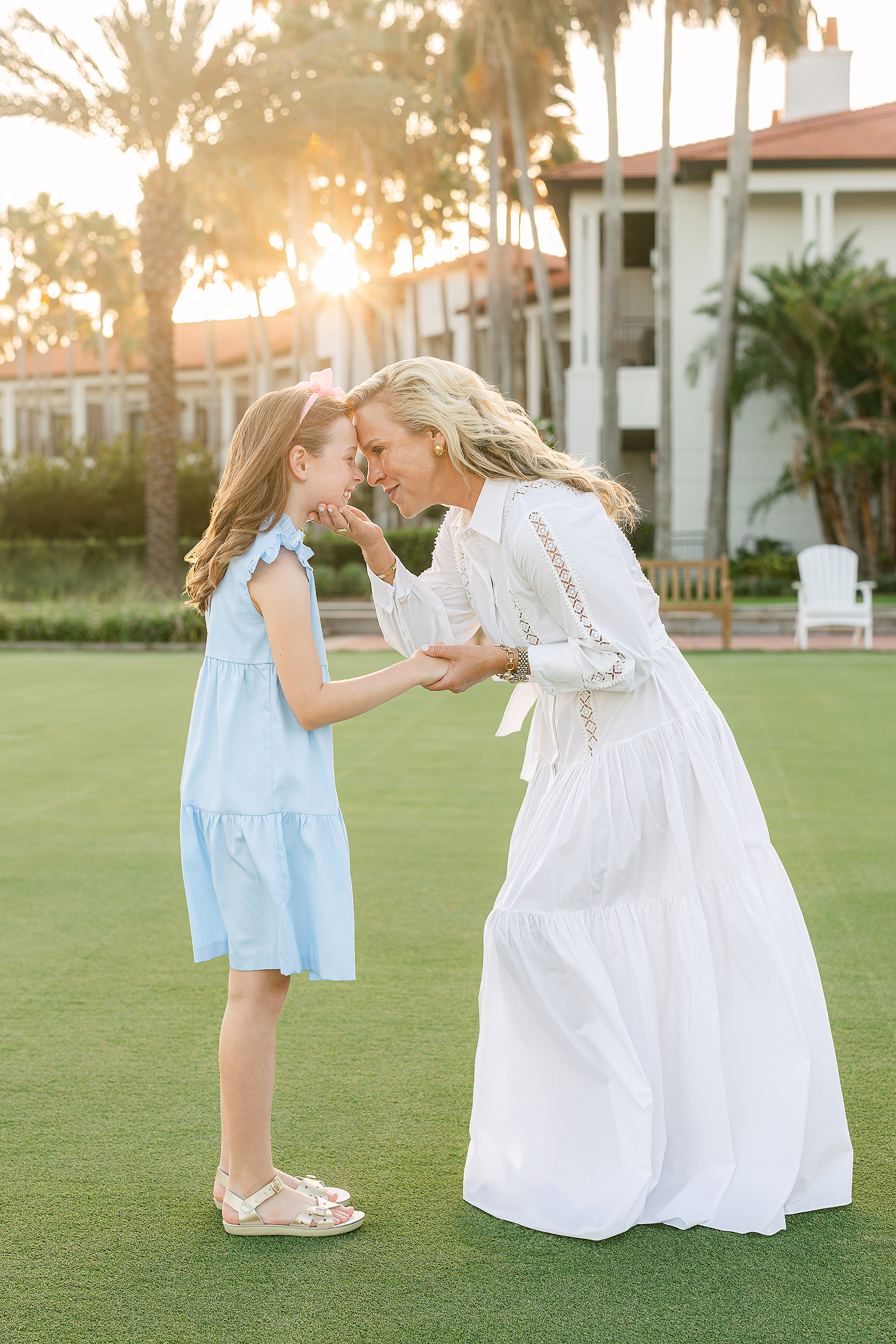 A woman in a white long dress hold her daughter's face at sunset on a golf course.