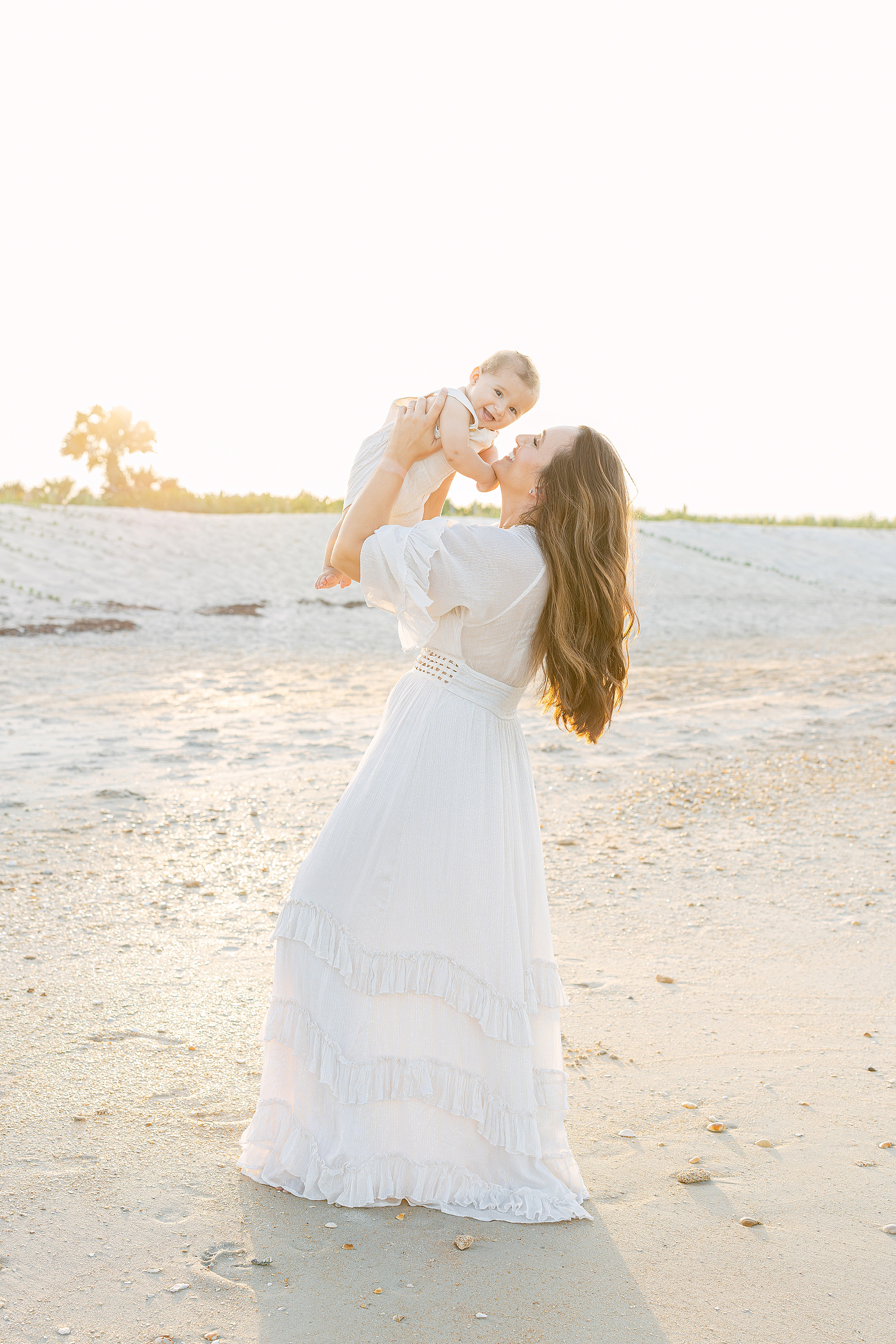 A woman in a white maxi dress holds up her baby boy on St. Augustine Beach at sunset.