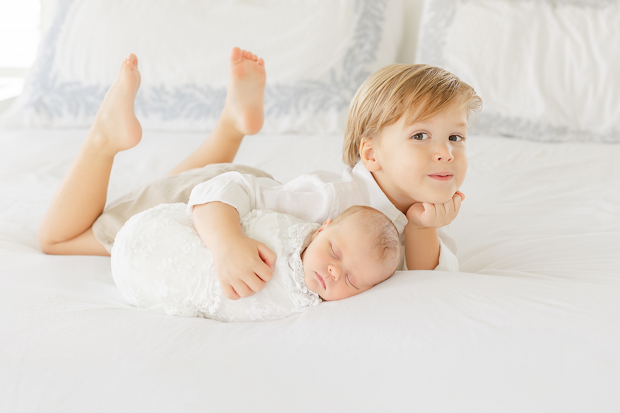 A toddler boy dressed in neutrals holds his newborn baby sister on a white bed at their home.