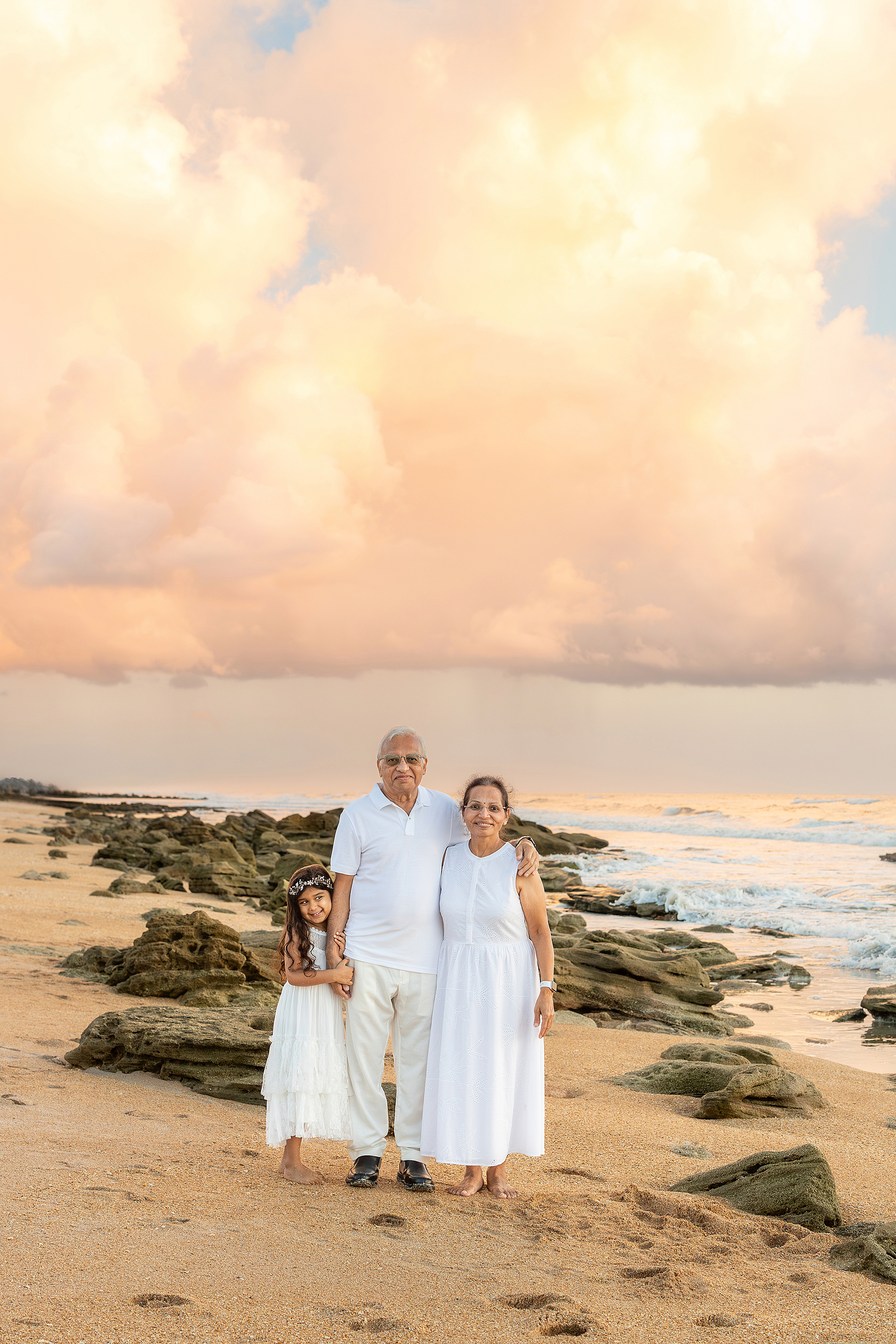 Grandparents stand with their granddaughter dressed in white on Saint Augustine Beach at sunrise.