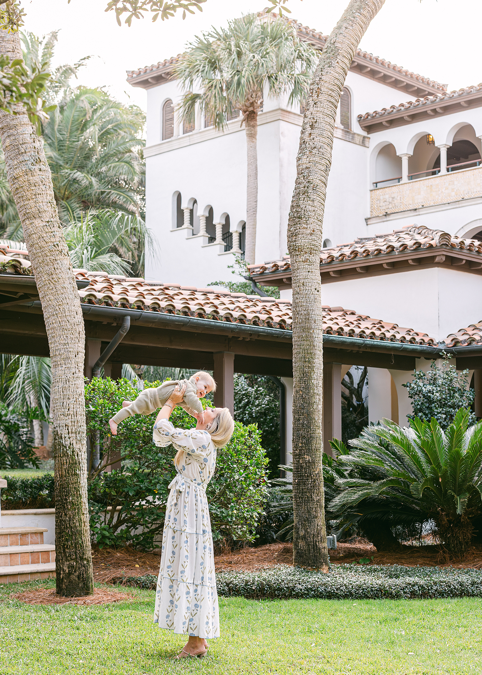 A woman in a blue and white maxi dress holds her baby boy high in the air on the grounds of the Cloister at Sea Island.