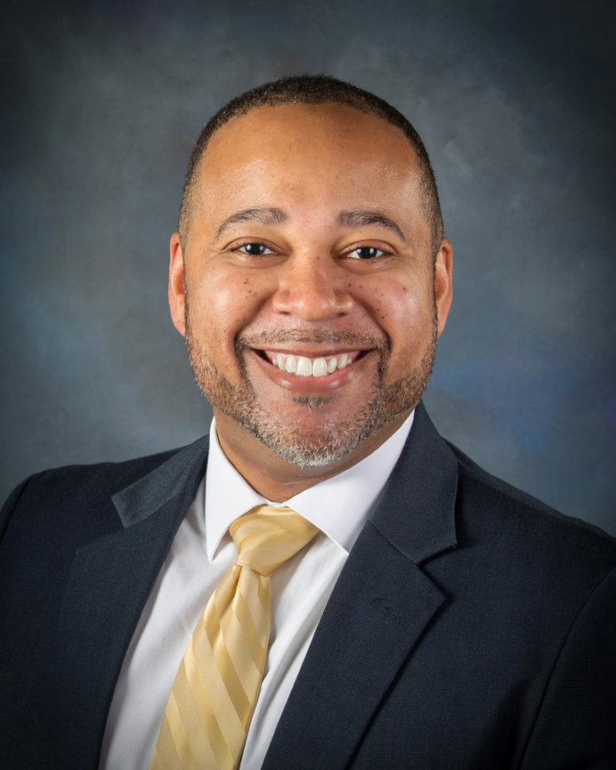 Smiling man in a dark suit with a yellow tie, against a gray background.