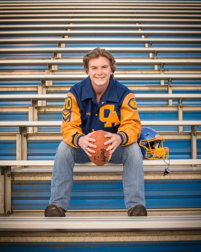 A person sits on bleachers, holding a football, with a sports jacket and helmet beside them.