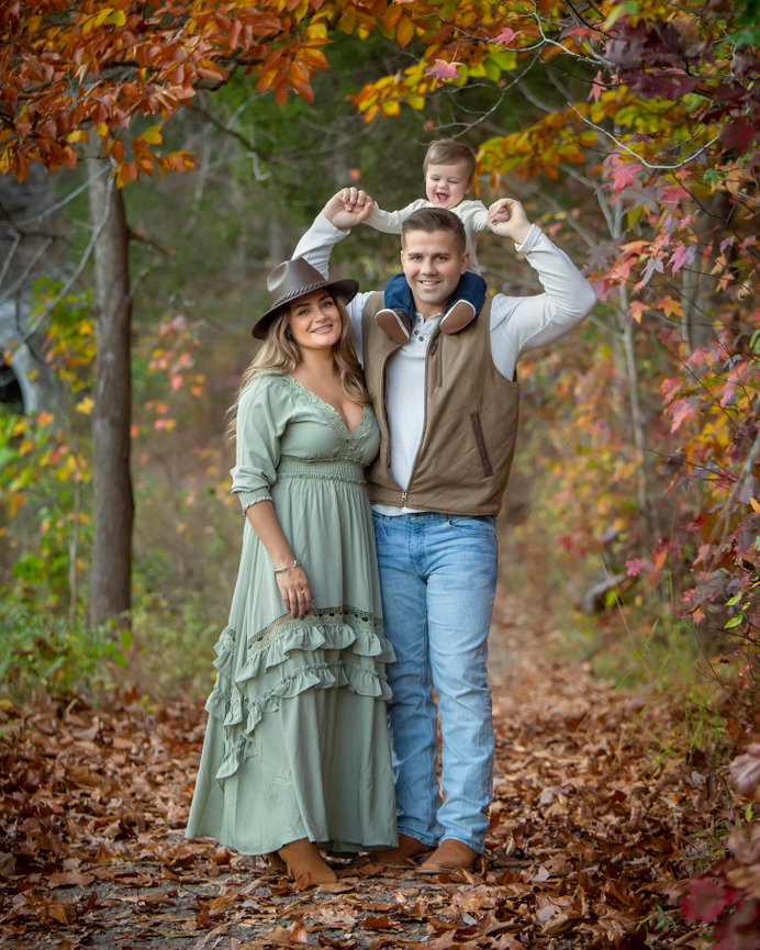 A family poses on a leaf-covered path, with vibrant autumn foliage in the background.