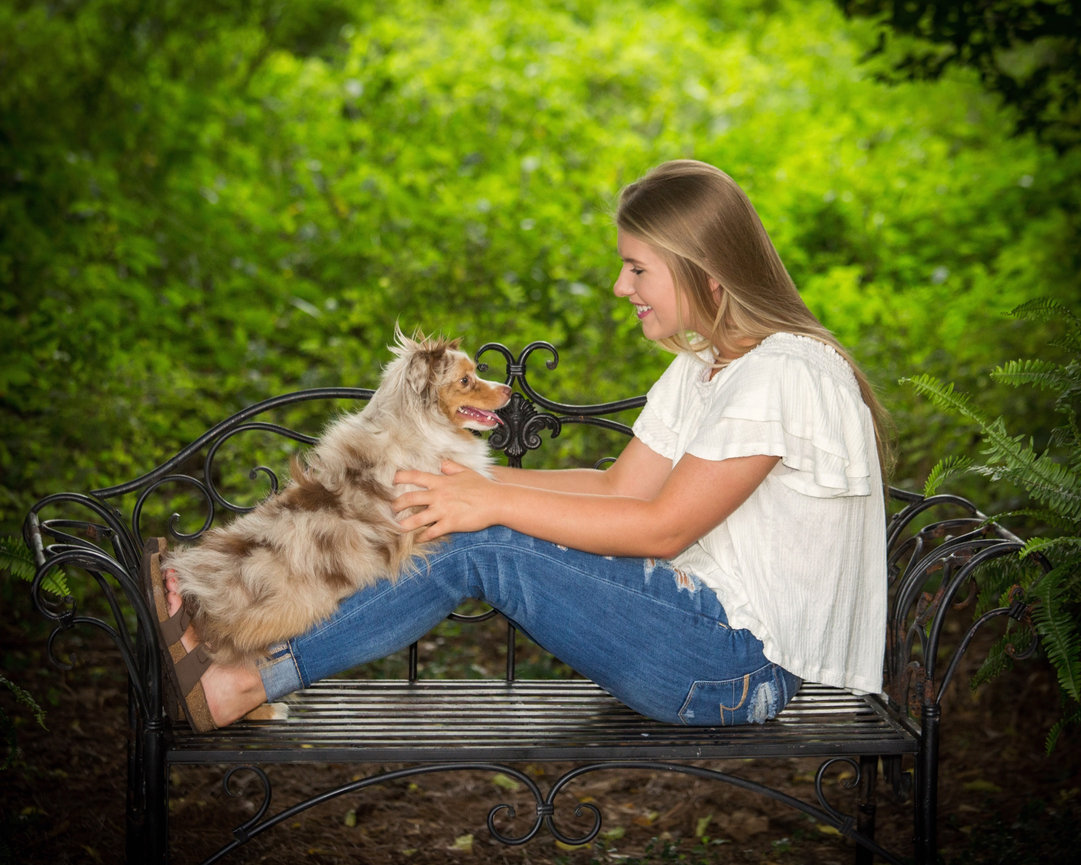 Woman sitting on a bench in a garden, smiling at a small dog on her lap.