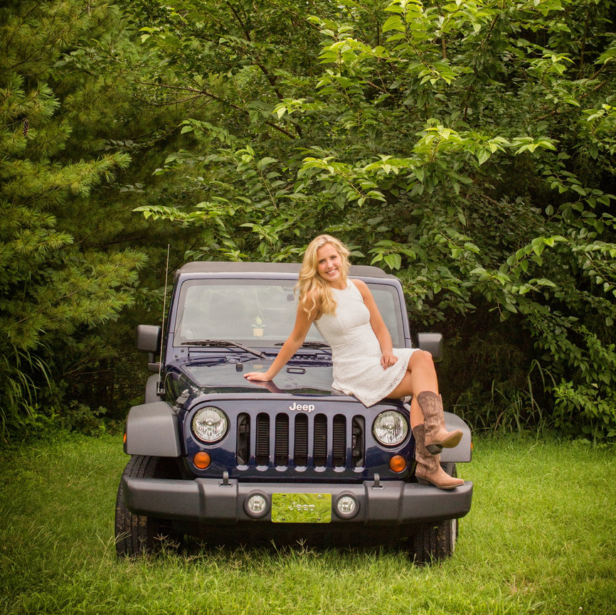 Woman in a white dress and boots sitting on a blue Jeep in a lush, green outdoor setting.
