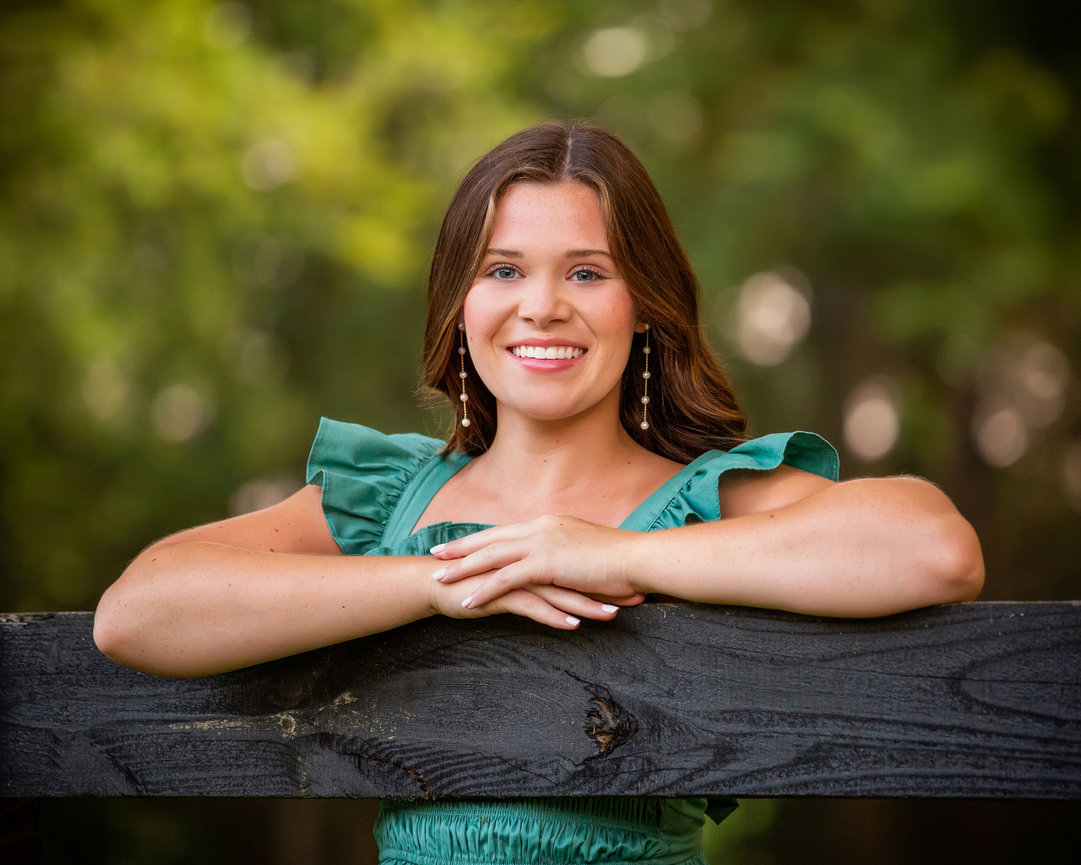 Smiling person in a green dress leans on a wooden fence with a blurred natural background.