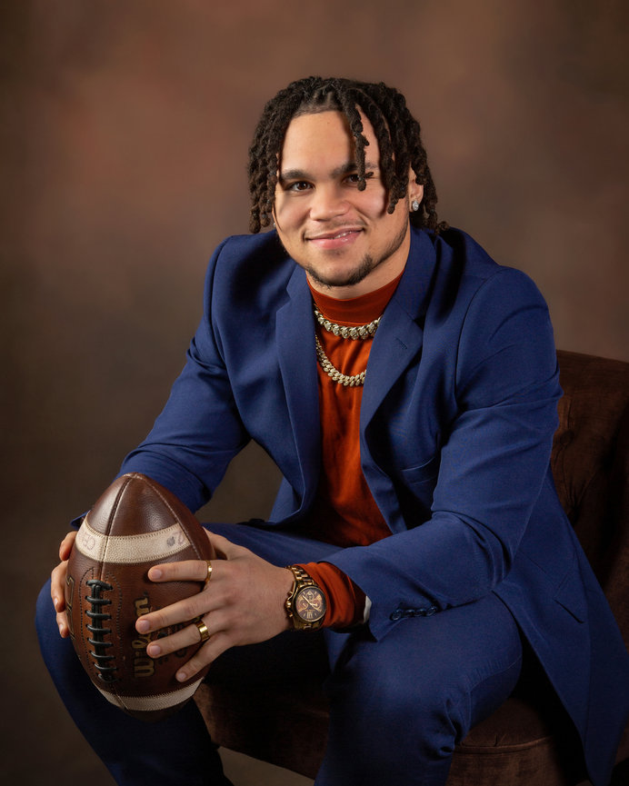 Man in a blue suit sitting with a football in hands against a brown backdrop.