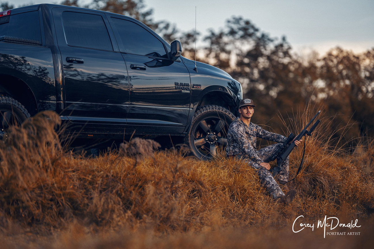 Man in camouflage holding rifle sits in front of black pickup truck on grassy terrain.