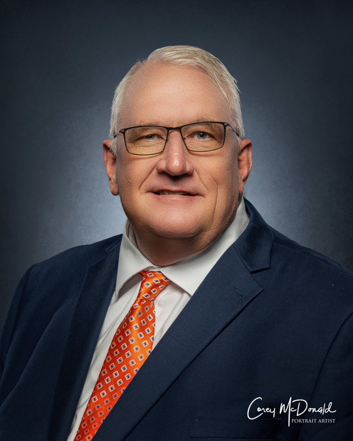 Older man in glasses and suit with an orange tie, studio portrait against a dark background.