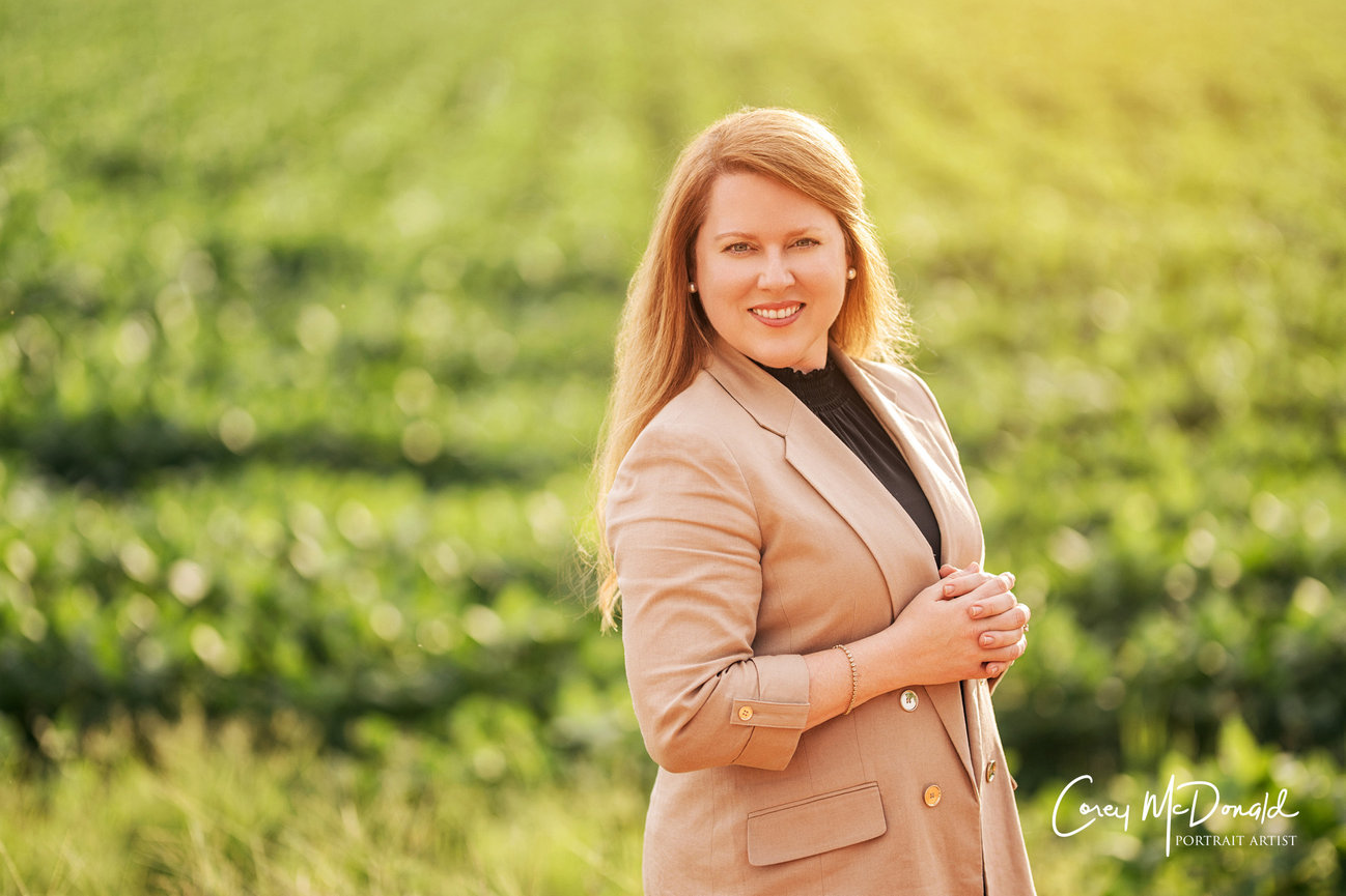 A person in a tan blazer stands in a lush green field, smiling warmly at the camera.