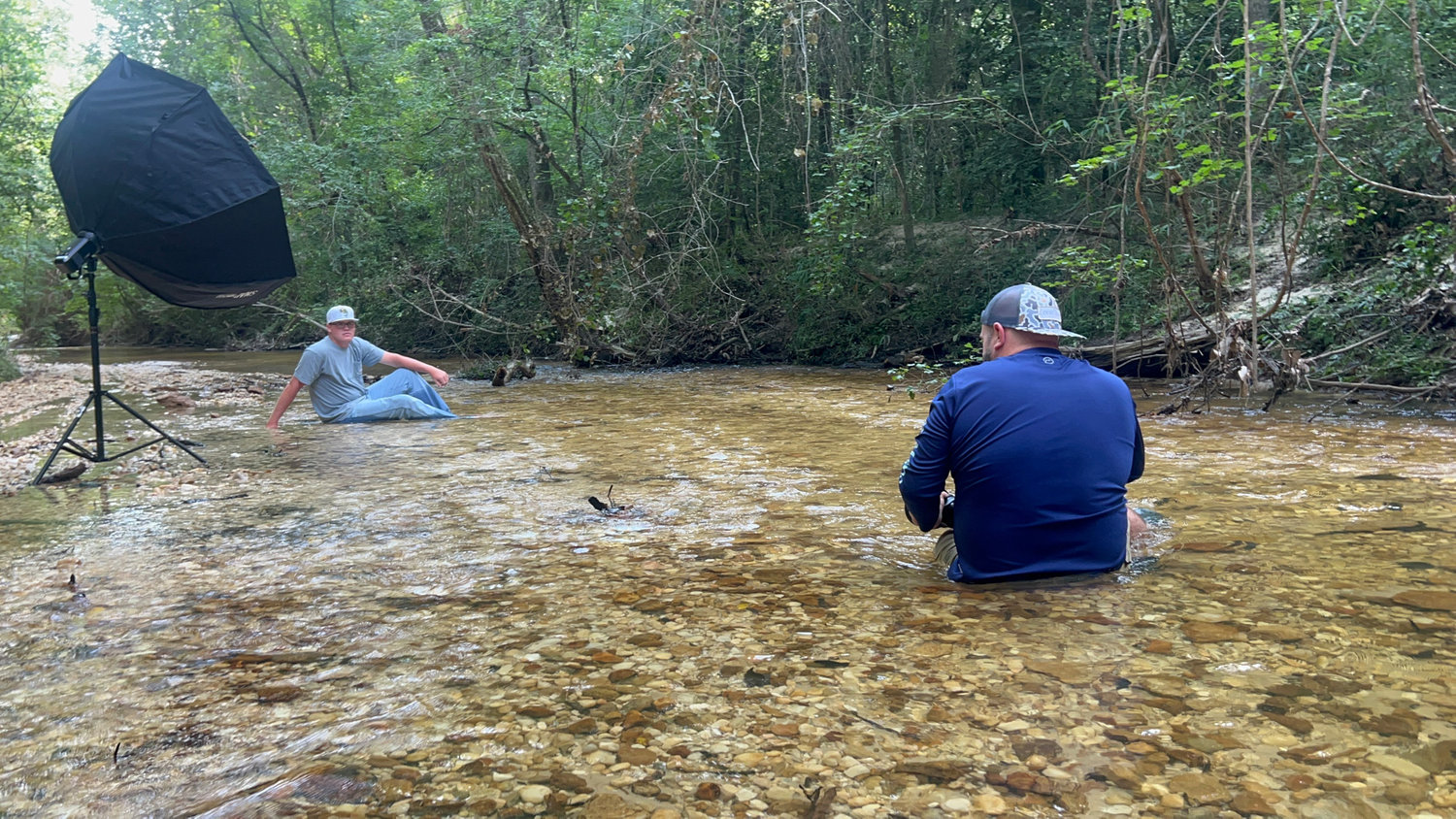 Two people sitting in a shallow creek, one with a lighting setup. Surrounded by trees in a natural setting.