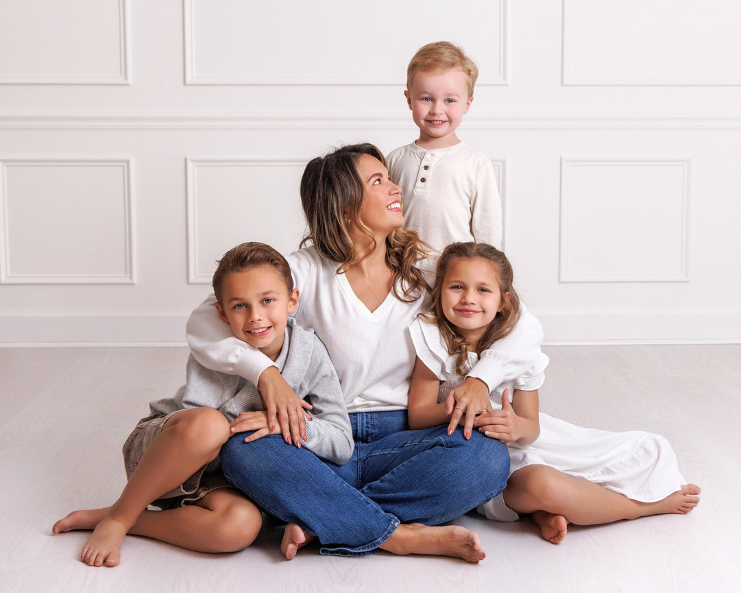 A woman sitting on the floor with three children, smiling and embracing each other, in a white room.