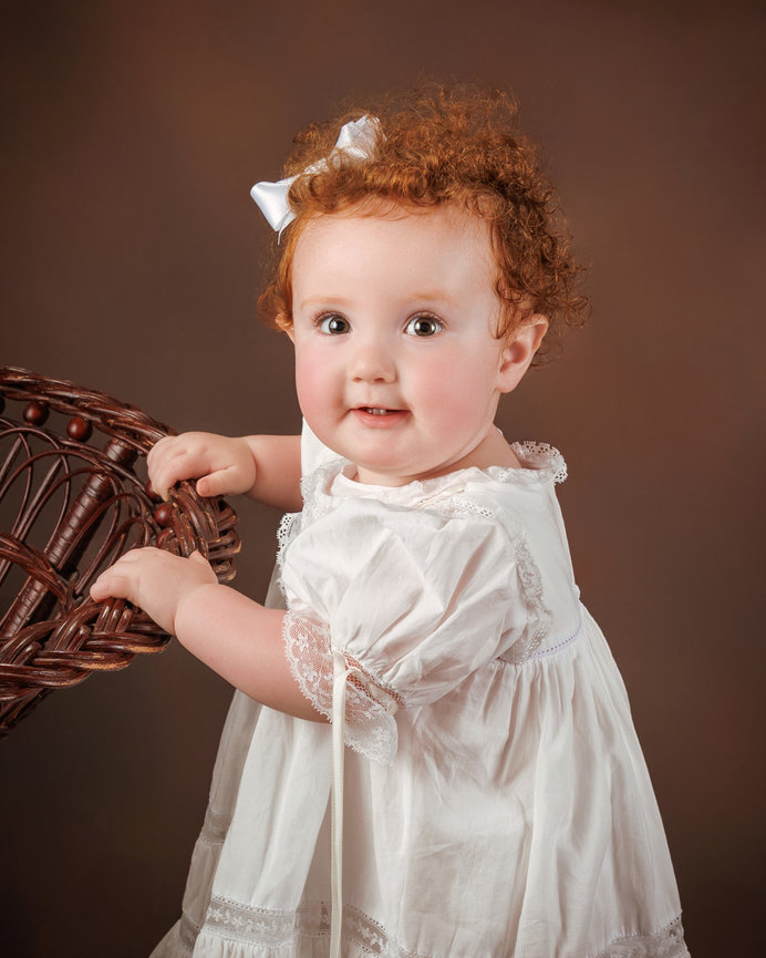 A toddler with curly red hair and a white dress holds onto a wicker chair against a brown background.