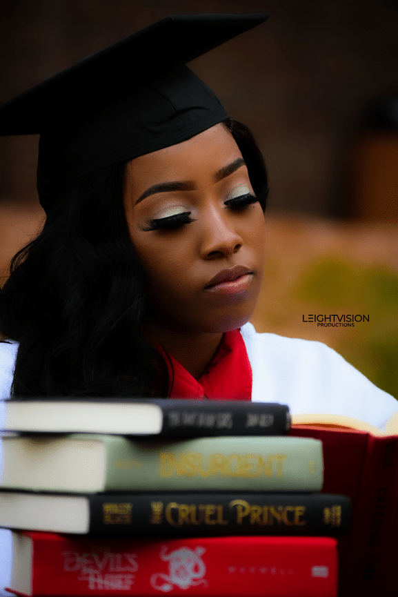 A black woman in a graduation cap and gown is focused on reading a book, symbolizing achievement and knowledge.