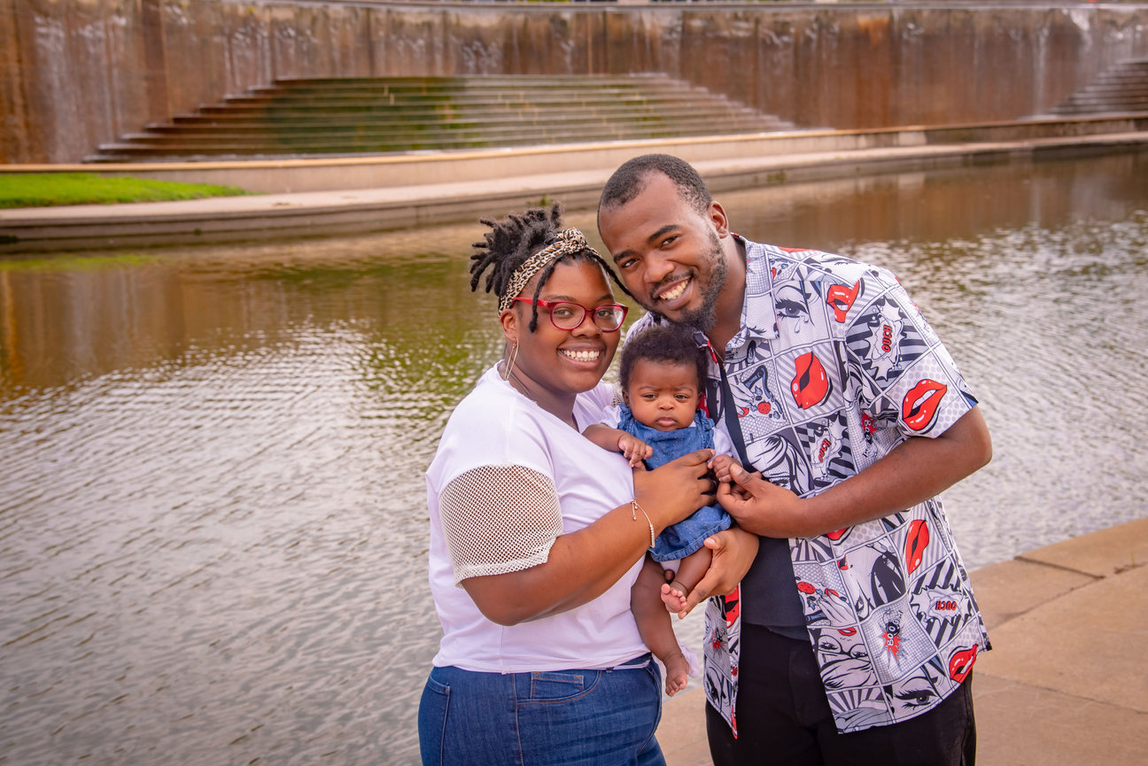 A happy family of three, smiling together by a water feature, with the mother holding a baby and the father beside them.