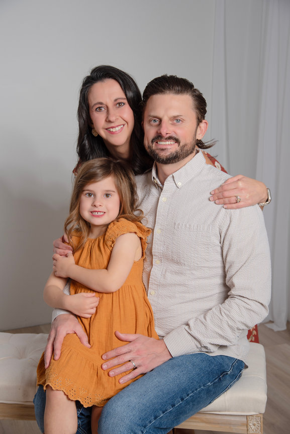 A family portrait featuring a smiling couple and their young daughter sitting together on a bench.