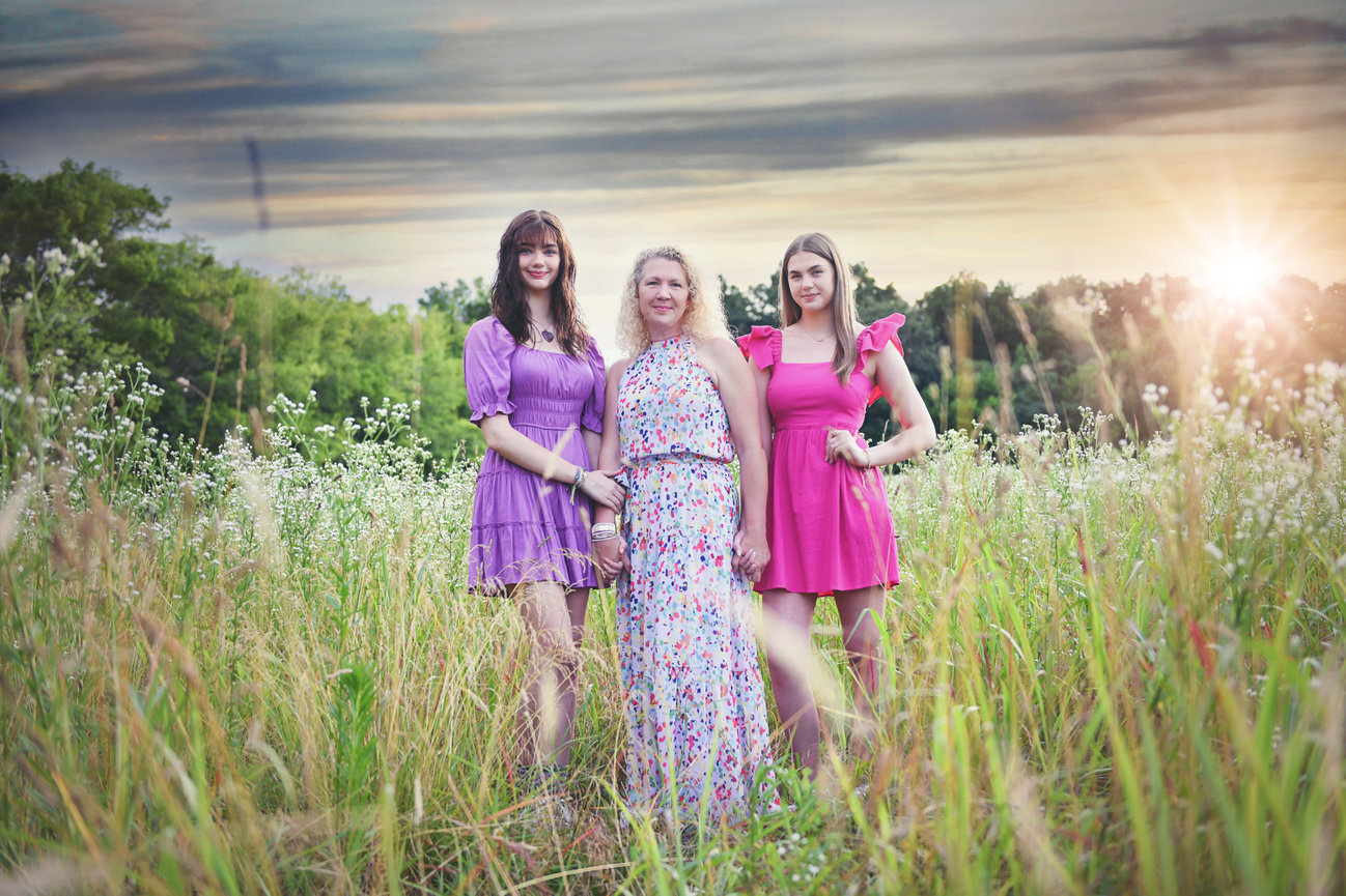Mom with two teenage daughters in pink and floral dresses standing in a tall wildflower field at sunset in Mocksville, NC during a family photo session