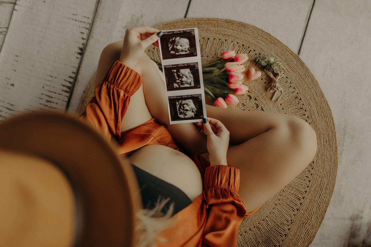 Pregnant woman holding ultrasound photos, seated on a round woven mat, with pink tulips beside her.