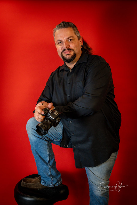 Man with a camera posing against a red backdrop, wearing a black shirt and jeans.