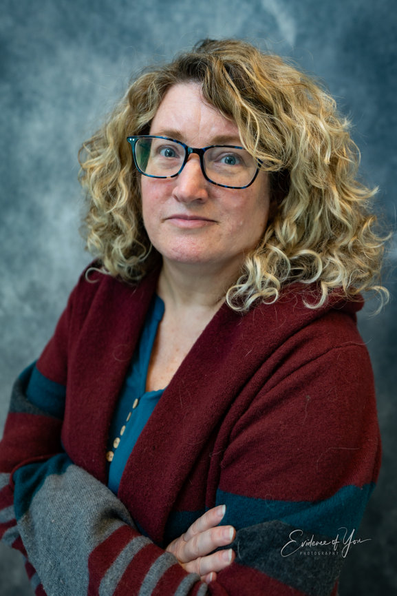A person with curly hair and glasses wearing a maroon and gray shawl against a gray backdrop.