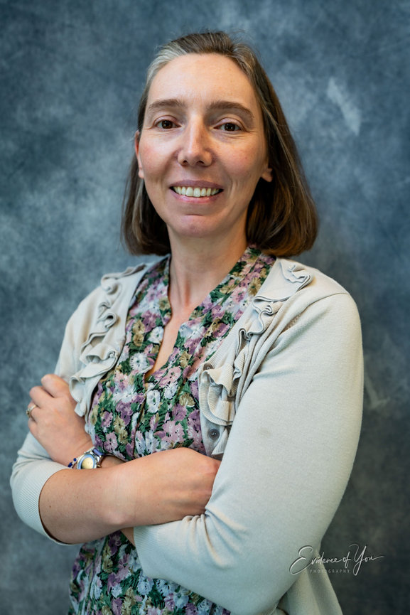 A person with short hair wearing a floral dress and beige cardigan, standing with arms crossed against a gray background.