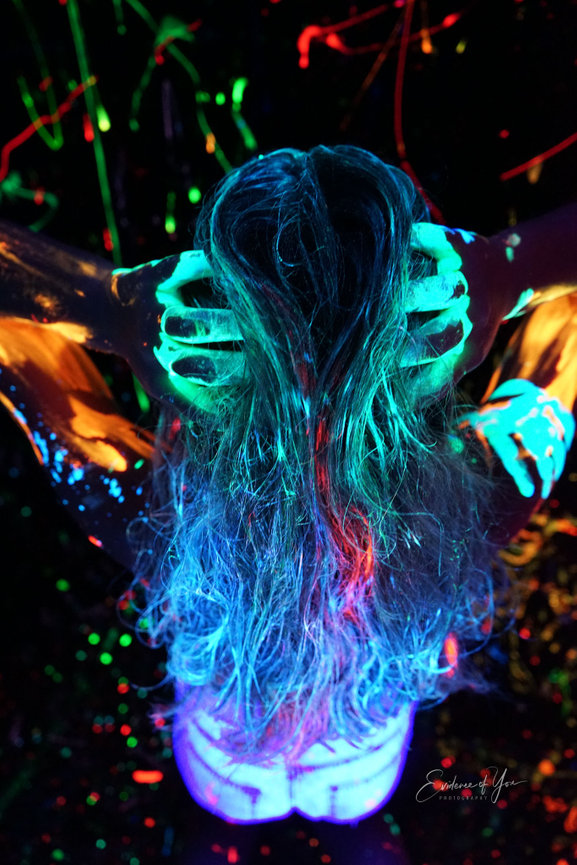 Person with neon body paint under UV light, holding their head, creating a vibrant, colorful glow effect.