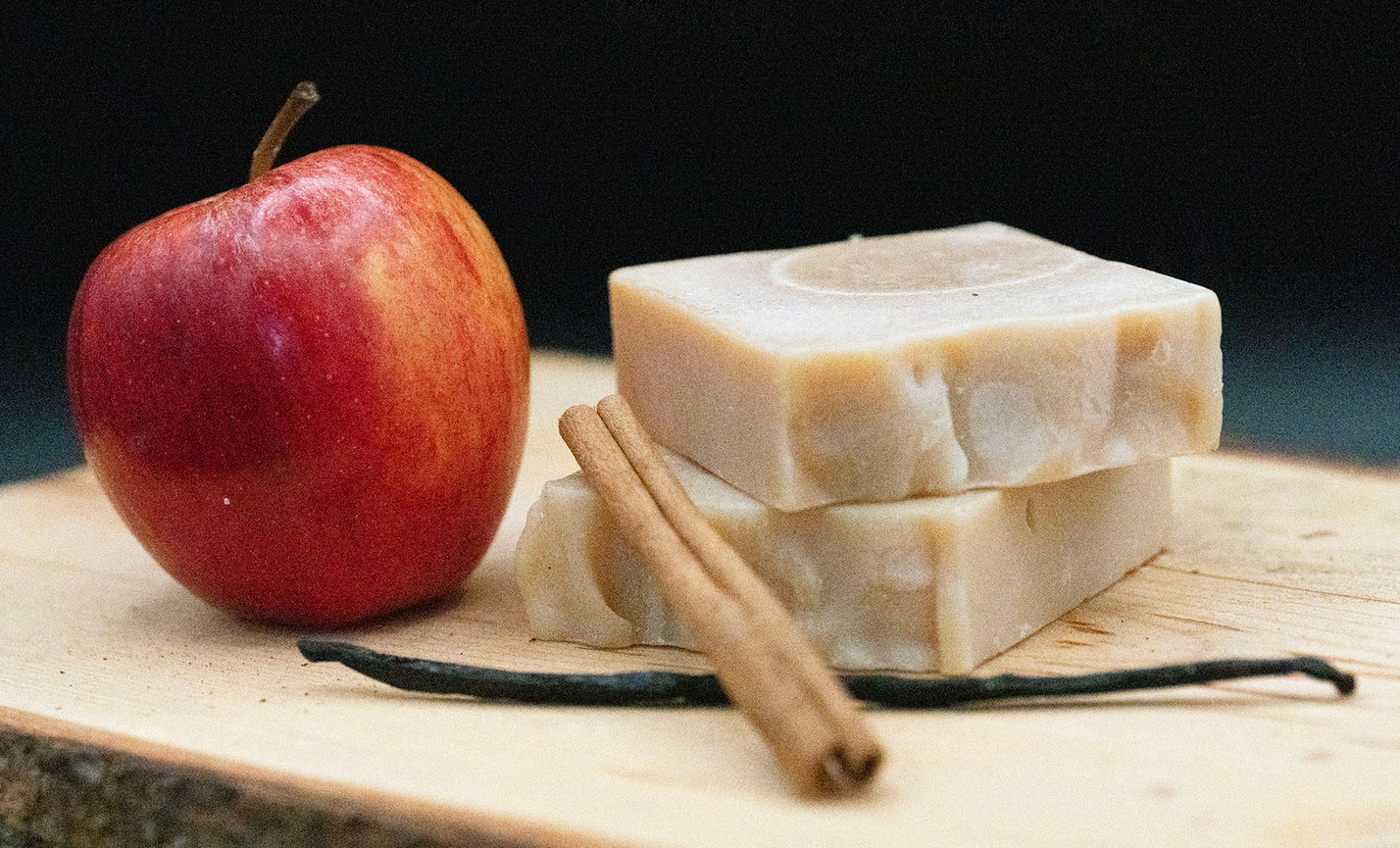 Apple, vanilla soap bars, cinnamon sticks, and a vanilla bean on a wooden board.