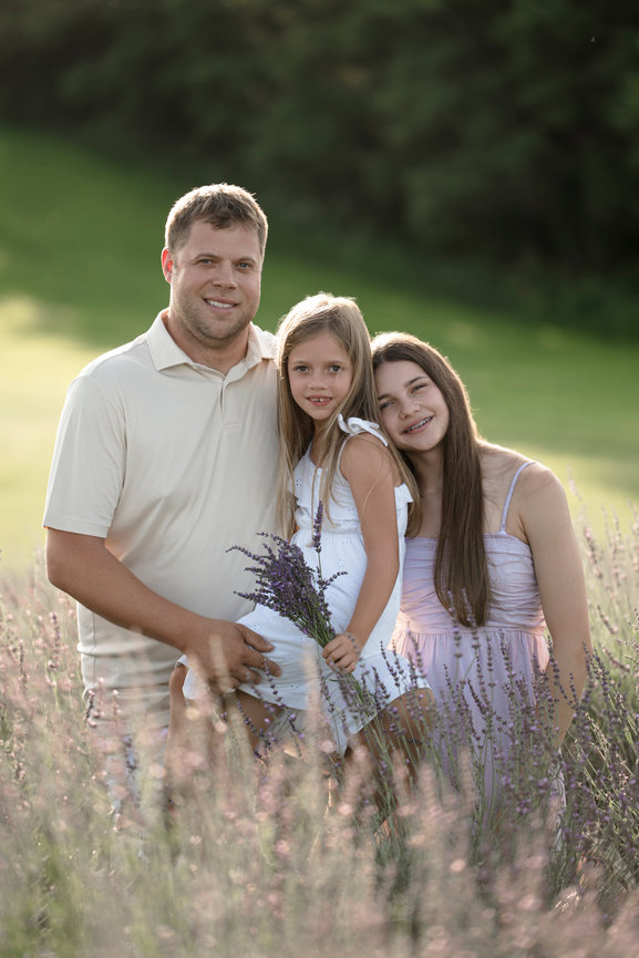 A father and two daughters stand together in a lavender field, smiling warmly at the camera.