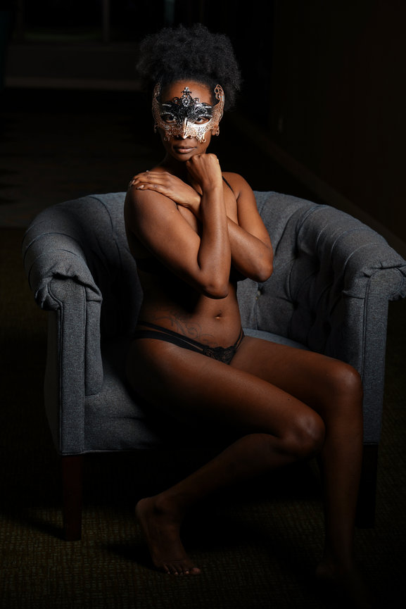 A seated individual with a decorative mask poses in a dimly lit space, highlighting their silhouette.