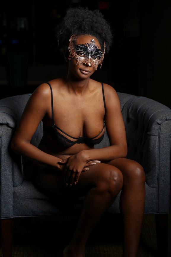 Person sitting in a chair wearing a decorative mask and lingerie, showcasing a moody atmosphere.