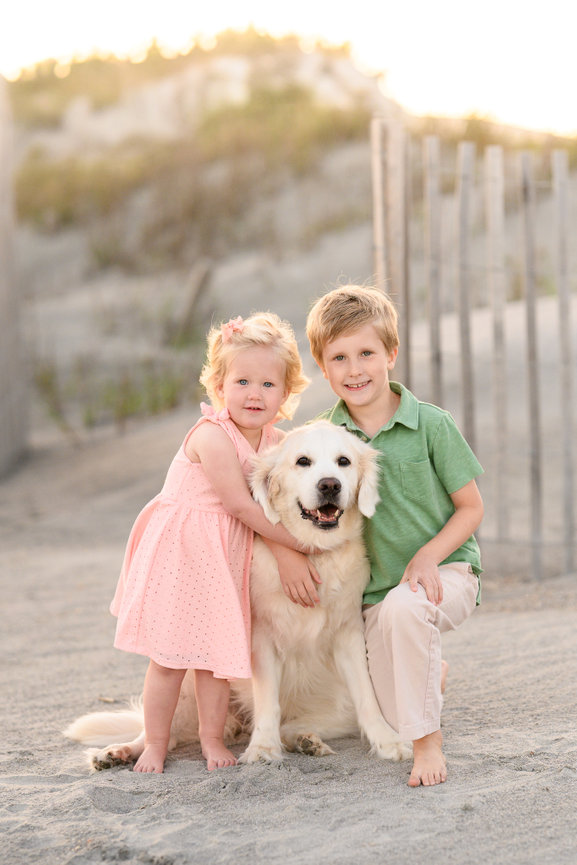 Two children smiling with a happy dog on a sandy beach, with dunes in the background.