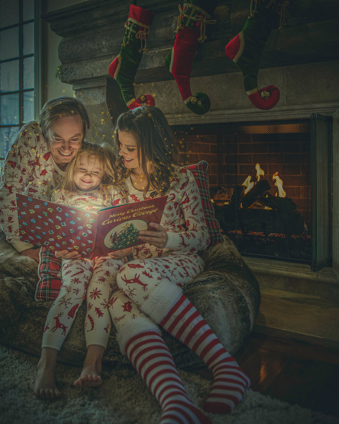 Family in matching pajamas reading a Christmas book by the fireplace, with stockings hanging above.