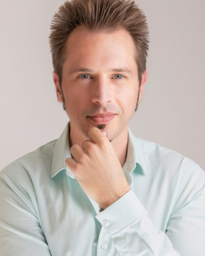 Man with light facial hair wearing a pale blue shirt, resting chin on hand, neutral background.