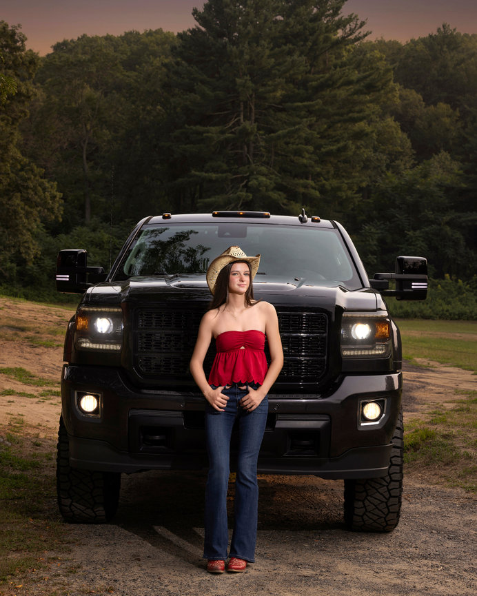 Woman in a red top and cowboy hat stands in front of a large black truck with headlights on, surrounded by trees.