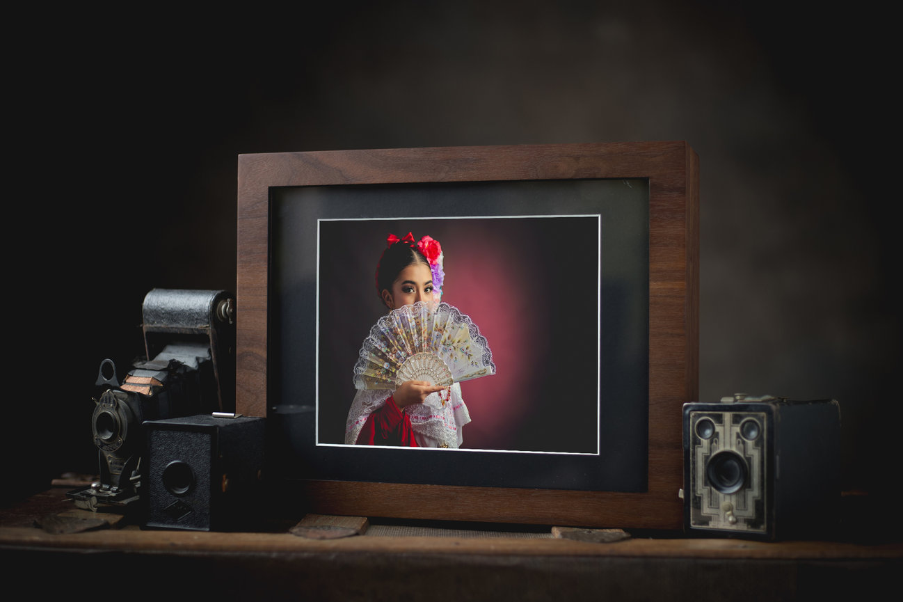 Framed portrait of a woman dressed in a traditional Mexican folkloric dress with a fan, surrounded by vintage cameras on a dark background.