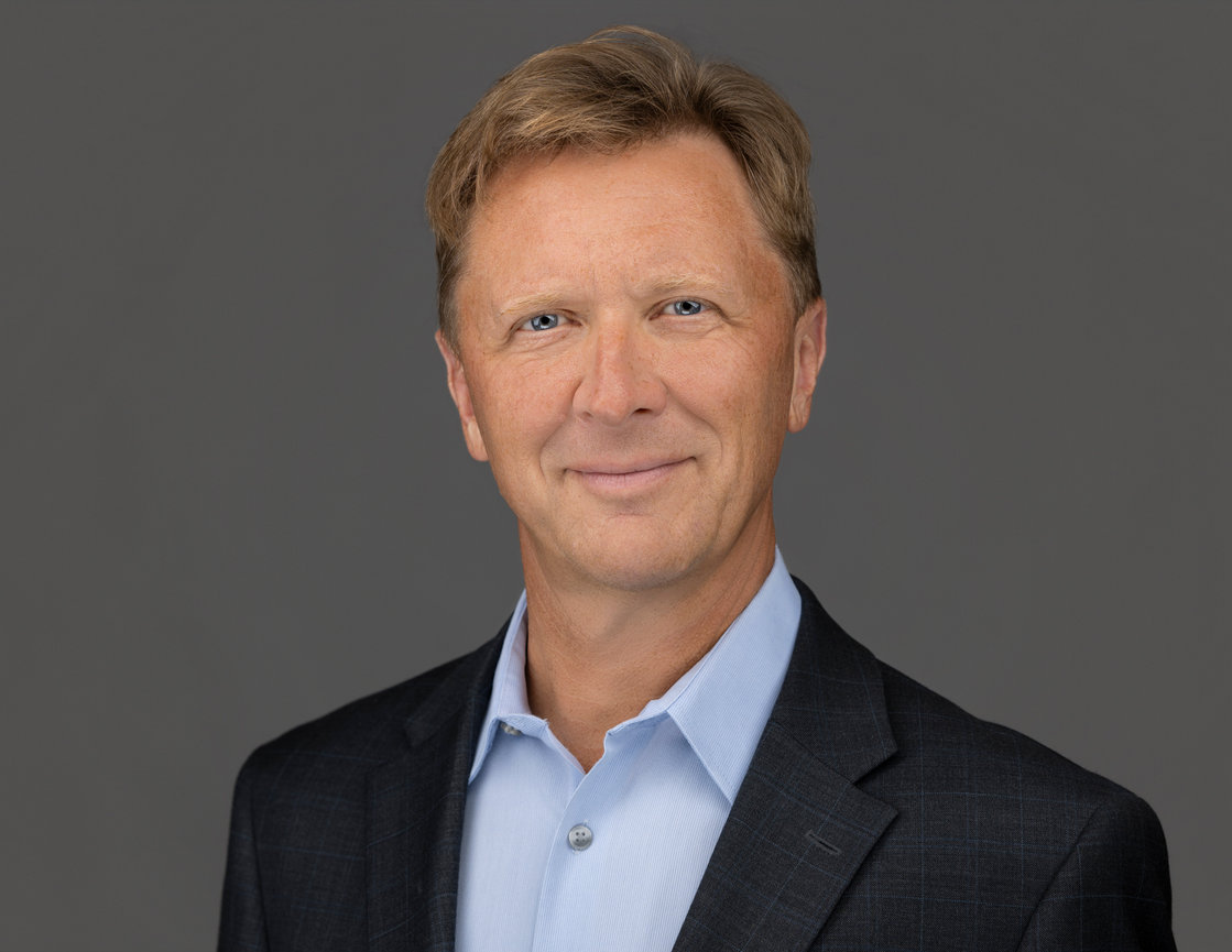 Portrait of a smiling man in a dark blazer and light blue shirt against a gray background