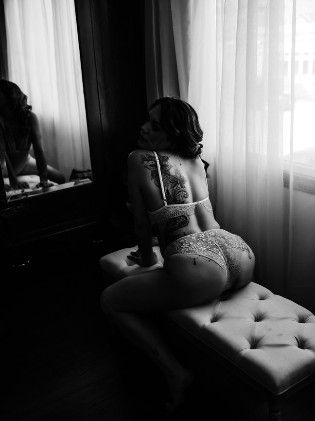 best nude boudoir photographer near me Port Angeles, WAPerson in lingerie with tattoos poses on a bench in a dimly lit room with curtains and a mirror.