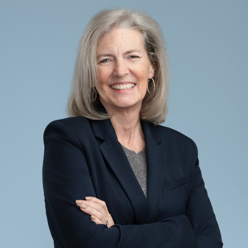 Smiling older woman in a dark blazer with arms crossed against a blue background