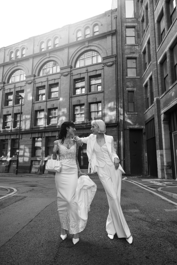 Editorial portrait of two brides—one in a tailored white trouser suit and the other in a long lace dress—posing on an empty Manchester street lined with historic brick buildings.