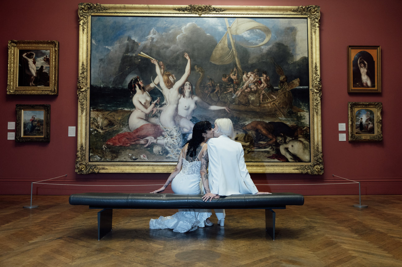 Direct flash editorial wedding photography of a same-sex couple in a white suit and lace dress, sitting before a grand framed painting at Manchester Art Gallery.