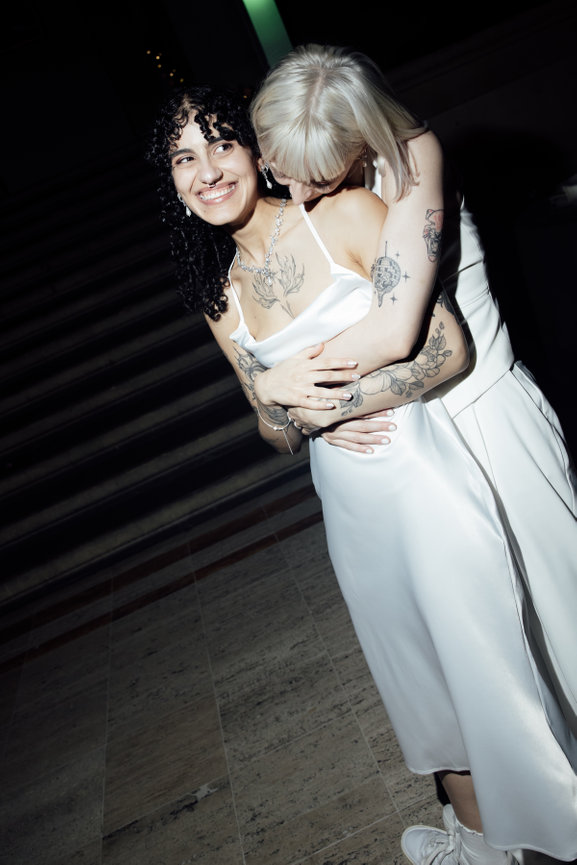 Editorial flash photography of two brides one in a white dress, one in a trouser suit, embrace, smiling, at Manchester art gallery at night.